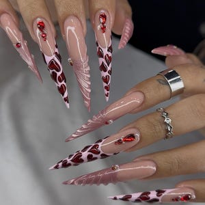 Op de afbeelding: Lange, puntige acrylnagels in een lichtroze tint, versierd met rode hartpatronen en sprankelende rode edelstenen. Sommige nagels hebben getextureerde, gebeeldhouwde details. Zilveren ringen zijn zichtbaar.