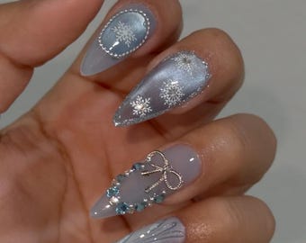 May include: Close-up of long, pointed fingernails with a winter theme. The nails are painted in shades of blue and silver, with snowflake and bow designs. Some nails have glitter accents and jewel embellishments.