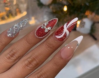 May include: A hand with long, pointed, artificial nails. The nails feature various designs, including silver glitter, red glitter with a bow, red glitter with a white design, and a French tip with rhinestones. The nails are a festive, holiday-themed style.
