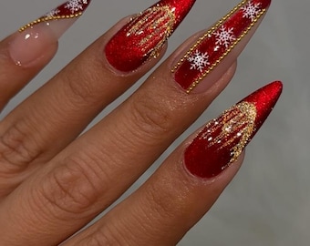 May include: Long, pointed fingernails with a festive design. The nails are painted a vibrant red with gold glitter accents and white snowflake details. A gold bead border adds a touch of elegance to the design, perfect for the holiday season.