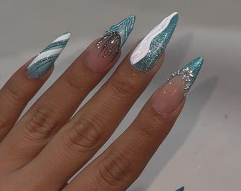 May include: A hand with long, pointed, teal-colored acrylic nails. Some nails feature glitter, white accents, and silver embellishments. One nail has a candy cane stripe design, while another has a silver glitter design near the cuticle.