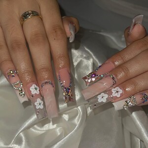 May include: Long, square-shaped acrylic nails with a light pink and white ombre design. Some nails feature white flower and heart-shaped rhinestone embellishments. Other nails are adorned with iridescent rhinestones along the edges.