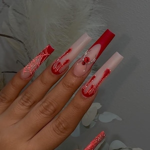 Pode incluir: Unhas acrílicas longas e quadradas com um design vermelho e transparente. Algumas unhas apresentam um acabamento com glitter vermelho, enquanto outras têm um design de chama vermelha. Uma unha tem um design de teia de aranha vermelha e outra um pingente de aranha prateada com pedras vermelhas.