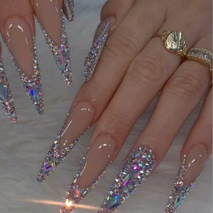 May include: Long, stiletto-shaped acrylic nails with a nude base and a sparkling, multi-colored rhinestone design. The rhinestones are arranged along the edges and tips of the nails, creating a glamorous and eye-catching look. Gold rings adorn the fingers.