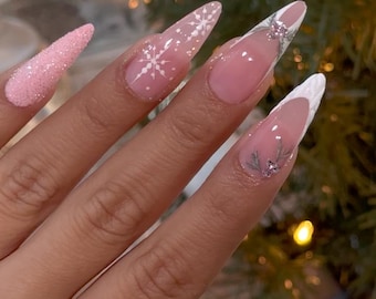 May include: Long, pointed fingernails with a winter theme. The nails feature a light pink base with white snowflake designs, glitter, and a white French tip. Some nails have small silver accents, creating a festive and elegant look.