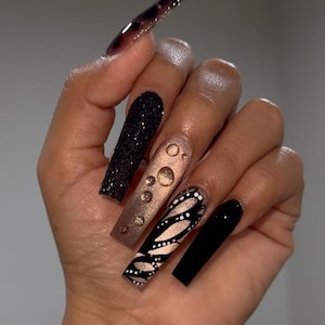 May include: A hand displaying long, coffin-shaped acrylic nails. The nails feature various designs, including black glitter, a gold and brown gradient, a butterfly pattern, and solid black. The nails are a mix of textures and colors.