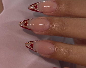 May include: Close-up of manicured nails with a red and gold design. The nails are oval-shaped with a clear base and a red tip. A gold glitter design is on the tip of each nail. The nails are on a light pink background.