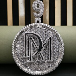 May include: A silver pendant with a circular face. The face displays the letters "DM" in a decorative font, encircled by a border of small, sparkling stones. The number "9" is at the top of the pendant, also set with stones.