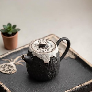 May include: A dark charcoal ceramic teapot with a textured, matte finish and a carved lotus pod motif. The lid features a matching lotus pod handle and a white, organic-shaped rim. The teapot has a curved spout and a sturdy, arched handle.