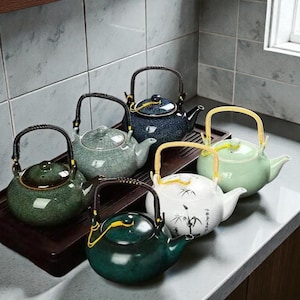 Puede incluir: Una colección de teteras de cerámica en varios colores, incluyendo verde, azul y blanco. Cada tetera tiene un asa y un pico. Algunas teteras tienen diseños decorativos. Las teteras están dispuestas sobre una bandeja de madera.