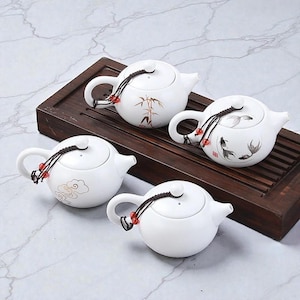 May include: Four white ceramic teapots with rounded bodies and curved handles are arranged on a dark wooden tray. Each teapot features a different decorative design, including bamboo, fish, and clouds. The teapots have a small, decorative cord with red beads.