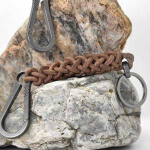 Op de afbeelding: Een bruin lederen gevlochten sleutelhanger met een metalen ring en twee metalen karabijnhaakclips. De sleutelhanger wordt getoond op een getextureerde grijze en bruine steen, met een witte achtergrond.