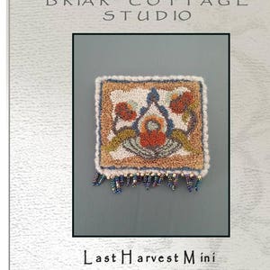 Puede incluir: Un patrón de punto de aguja o de bordado para una alfombra pequeña o un tapiz de pared. El diseño presenta un motivo floral estilizado en tonos naranja, marrón y verde, con un borde de flecos de cuentas. El patrón se titula "Last Harvest Mini" y es de Briar Cottage Studio.