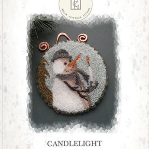 Puede incluir: Un adorno de punto de aguja con un muñeco de nieve sosteniendo una vela. El adorno está enmarcado en un aro de alambre de cobre y tiene un fondo azul y blanco con detalles marrones. El texto "CANDLELIGHT" está impreso debajo del adorno.