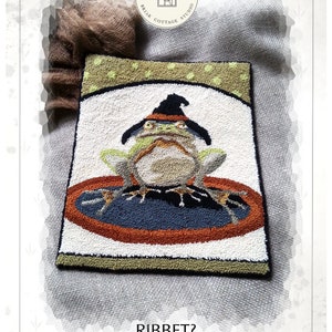 Peut inclure: Un modèle de point de broderie représentant une grenouille verte portant un chapeau de sorcière et assise sur un cercle noir et orange. Le modèle est intitulé "Ribbet?" et est conçu par Kate Gillery.