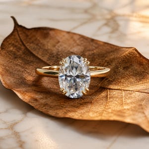 Puede incluir: Un anillo de compromiso de oro con un diamante de talla ovalada. El anillo está sobre una hoja marrón, con el diamante reflejando la luz. La banda es delgada y simple, y las garras que sujetan el diamante son visibles.