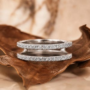 May include: A silver-toned double band ring featuring two parallel rows of small, clear, round-cut stones set along the entire circumference of each band, connected by a central bridge, creating a stacked appearance with a polished finish.