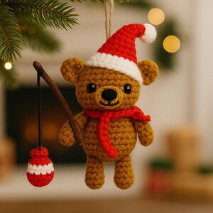 May include: Crocheted Christmas ornament of a brown bear wearing a red Santa hat and scarf. The bear holds a fishing rod with a red and white striped bobber. The ornament is hanging from a green tree branch.