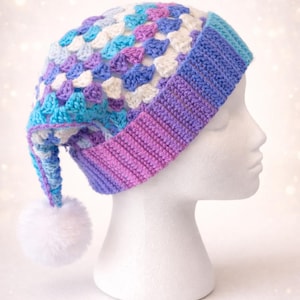 May include: A handmade crochet hat in a colorful patchwork design. The hat features a mix of blue, purple, and white squares, with a ribbed brim that transitions from purple to blue. A white pom-pom dangles from the end.