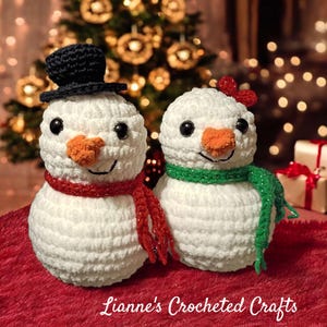 May include: Two handmade crochet snowmen. One snowman wears a black top hat and a red scarf, the other has a red bow and a green scarf. Both have orange carrot noses, black eyes, and smiling mouths. The snowmen are on a red surface.