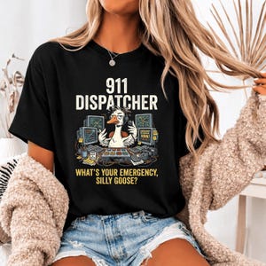 May include: Black t-shirt with the text "911 DISPATCHER" above a cartoon goose wearing a headset and sitting at a computer. The phrase "WHAT'S YOUR EMERGENCY, SILLY GOOSE?" is printed below the goose.