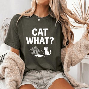 Könnte beinhalten: Dunkelgrünes T-Shirt mit dem weißen Text "CAT WHAT?". Darunter eine Grafik eines umgestürzten Blumentopfs, einer Pflanze und einer Katze. Das T-Shirt hat kurze Ärmel.