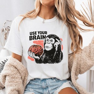 May include: White t-shirt with a black and white illustration of a chimpanzee holding a brain, with the text "USE YOUR BRAIN" above the image. The brain is red and the background is a light red color.