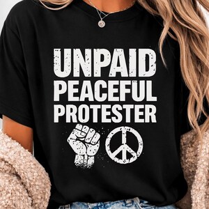 May include: Black t-shirt featuring the white text "UNPAID PEACEFUL PROTESTER", a white clenched fist, and a peace symbol. A casual item of clothing.