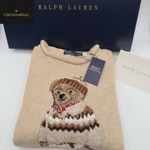 May include: A beige Polo Ralph Lauren sweater featuring a bear design. The bear is wearing a hat and scarf. The sweater has a crew neck and long sleeves. A Polo by Ralph Lauren tag is attached. The Ralph Lauren box is in the background.