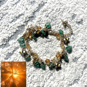 May include: A delicate bracelet with a nature theme. The bracelet features green leaf charms, small yellow bee charms, and amber-colored crystal beads. The bracelet is made of silver-colored metal and is displayed on a textured white surface.