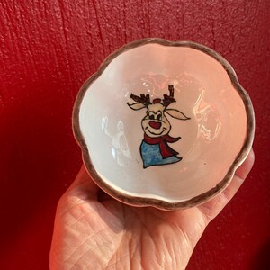 May include: A small, white ceramic bowl with a scalloped edge and a brown rim. The bowl features a hand-painted cartoon reindeer with a red nose, scarf, and hat. The bowl is held against a red background.