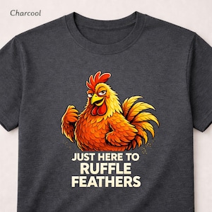 May include: A charcoal grey t-shirt with a cartoon rooster design. The rooster is depicted with orange and yellow plumage and a red comb. The text "JUST HERE TO RUFFLE FEATHERS" is printed below the rooster.