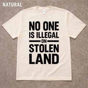 May include: Cream-colored t-shirt with the text "NO ONE IS ILLEGAL ON STOLEN LAND" in bold, black letters. The shirt has a crew neck and short sleeves. The word "NATURAL" is in the top left corner.