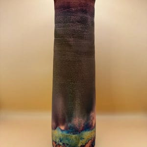 May include: A tall, handcrafted ceramic vase with a flared top. The vase features a gradient of colors, transitioning from a deep red at the top to shades of purple, brown, and teal at the base. The textured surface adds depth to the design.