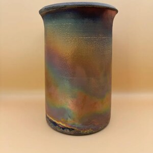 Könnte beinhalten: Eine zylindrische Keramikvase mit ausgestelltem Rand. Die Vase hat eine metallische Oberfläche mit schillernden Farben von Blau, Grün, Gold und Kupfer. Der Boden der Vase hat einen dunkelblauen Farbton.