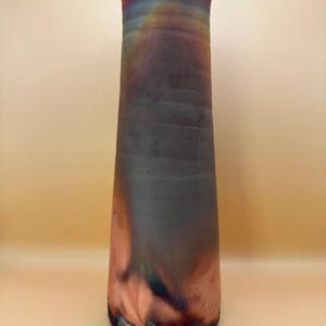 May include: A tall, iridescent ceramic vase with a flared top. The vase transitions from a dark gray to a reddish-brown hue, with hints of blue and green. The vase is approximately 12 inches tall.