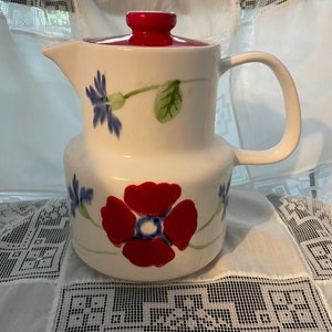 May include: White ceramic pitcher with a red lid and floral design. The flowers are red, blue, and green.