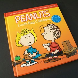 May include: A hardcover cookbook titled "PEANUTS Lunch Bag Cookbook" with illustrations of Peanuts characters. The cover is orange and yellow with the title in large, bold letters. The book features over 50 packable snacks and sandwiches.