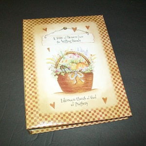 May include: A decorative book with a floral basket illustration. The cover features a brown and tan checkered border and the text "A Taste of Heaven from the Willing Hands" and "Lakemoore Church of God of Prophecy".