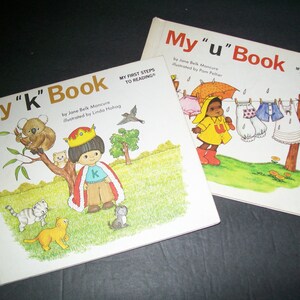 May include: Two children's books, "My "k" Book" and "My "u" Book", both part of the "My First Steps to Reading" series. The "k" book features a child dressed as a king with a koala and kittens. The "u" book features a child in a yellow raincoat holding an umbrella with clothes hanging on a line.
