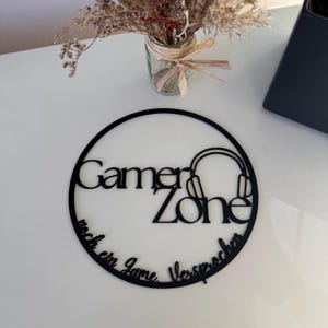 Könnte beinhalten: Schwarzes Gamer Zone-Schild mit kreisförmigem Design. Das Schild zeigt die Wörter "Gamer Zone" und "noch ein Game versprochen" mit einer Kopfhörergrafik. Das Schild befindet sich auf einer weißen Oberfläche.