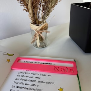 Könnte beinhalten: Ein leuchtend pinkfarbenes, rechteckiges Lese-Lineal mit einem Sichtfenster und dem eingeprägten Namen Nala auf der rechten Seite. Das Lese-Lineal liegt horizontal auf einer offenen Buchseite mit schwarzem gedrucktem Text.
