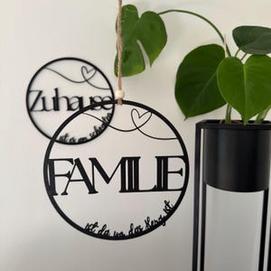 Könnte beinhalten: Schwarzes Metall-Familienschild mit dem Wort „FAMILIE“ in großen Buchstaben und dem Satz „ist da wo das Herz ist“ in kleinerer Schrift. Ein zweites Schild mit dem Wort „Zuhause“ ist ebenfalls sichtbar.
