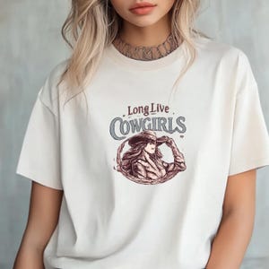 May include: Cream-colored t-shirt featuring the text "Long Live Cowgirls" above a graphic of a cowgirl in a hat. The illustration is in shades of brown and red. The shirt has short sleeves and a crew neck.