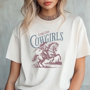 May include: Cream-colored t-shirt featuring a graphic of a cowgirl riding a horse, with the words "Long Live Cowgirls" in blue and white. The design is a vintage-style illustration, perfect for fans of Western themes.