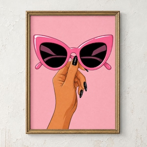 Könnte beinhalten: Ein gerahmter Kunstdruck, der eine Hand mit schwarzem Nagellack zeigt, die eine leuchtend rosa Katzenaugen-Sonnenbrille mit dunklen Gläsern vor einem einfarbigen hellrosa Hintergrund hält. Der Rahmen ist goldfarben und hat eine strukturierte Oberfläche.