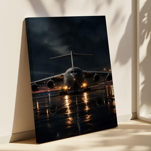 Könnte beinhalten: Ein großes militärisches Transportflugzeug steht nachts auf einem nassen, reflektierenden Rollfeld. Das dunkelgraue Flugzeug hat eingeschaltete Landescheinwerfer, die einen warmen Schein auf den Boden werfen. Der Himmel ist dunkel und bewölkt.
