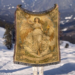 May include: A woven tapestry blanket featuring a classical illustration of a winged figure holding a laurel wreath and a torch. The blanket has a beige and brown color palette with the words "VICTORIA + GLORIA" on a banner. The blanket has a fringed edge.
