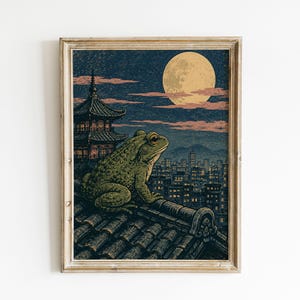 Könnte beinhalten: Ein gerahmter Kunstdruck, der einen großen grünen Frosch auf einem dunklen Ziegeldach zeigt. Im Hintergrund beleuchtet ein Vollmond eine Stadtlandschaft mit einer Pagode unter einem dunkelblauen Himmel mit zartrosa Wolken, alles in einem hellen Holzrahmen.
