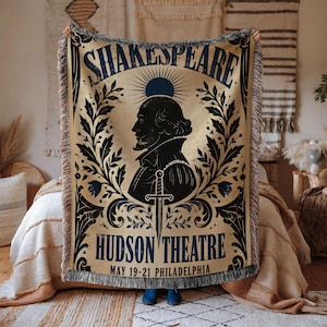 May include: A woven throw blanket featuring a black silhouette of William Shakespeare centered within a dark blue floral frame. The text reads SHAKESPEARE, HUDSON THEATRE, and MAY 19-21 PHILADELPHIA. The blanket has a fringed edge and tan background.